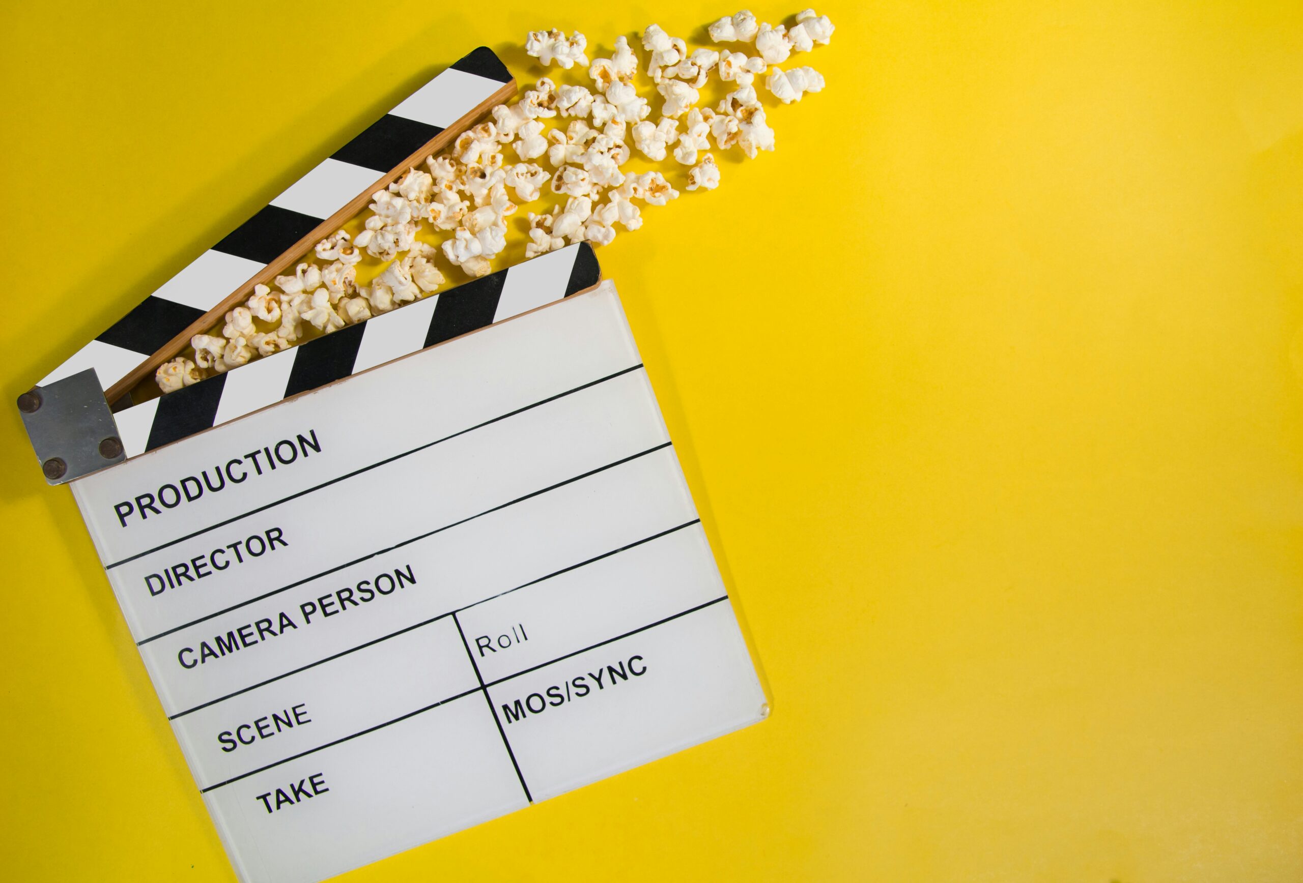 Black and white film clapper board on bright yellow background. with popcorn spilling from its "mouth"